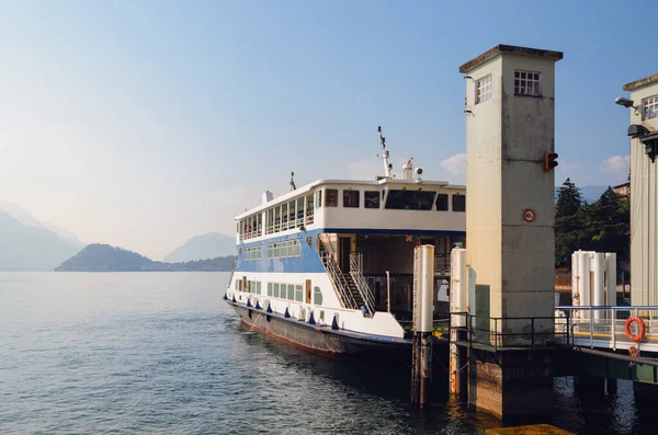 Sabahın erken saatlerinde, Menaggio, İtalya limanına yanaşmış bir feribot ile Como Gölü manzarası ve açık gökyüzü, sis, güneş ışığı ve dağlarla birlikte.