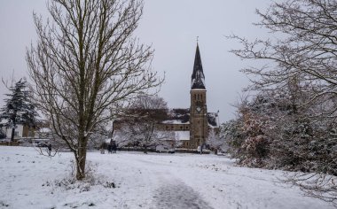 Woodford Green 'deki All Saints Kilisesi' nin karla kaplı zemin ve ağaçlarla dolu sakin kış sahnesi. Kış, inanç, cemaat, miras ve mevsimsel manzaralar temaları için uygun sakin ve atmosferik bir görüntü..