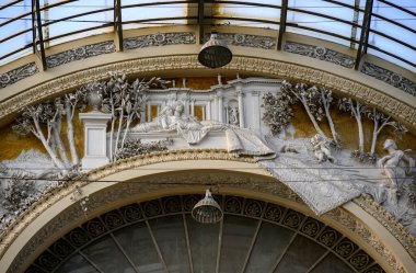 Napoli 'deki Galleria Umberto' nun iç mimari ve dekoratif unsurlarını içeren iç detayları. Süslü friz klasik figürler ve levrek güvenlikli ayrıntılı ağaçlar tasvir ediyor. Kemerli yapıda metal çerçeveli cam bir çatı yer alıyor.