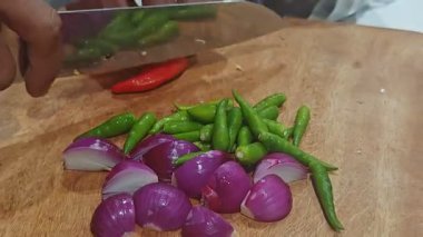 Kırsal bir ahşap kesim tahtasındaki taze yeşil kuş gözü biberlerini (Tayland biberleri) kesmek için paslanmaz çelik mutfak bıçağı kullanan birinin yakın plan fotoğrafı. Ön planda dilimlenmiş kırmızı soğanlar görülebiliyor, ev yemeklerini gösteriyor..