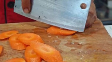 Bir şefin elleri taze, parlak turuncu havuçları ince ince dilimlemek için büyük paslanmaz çelik bir satır kullanır. Video profesyonel bıçak becerilerini ve sebzelerin canlı dokusunu ön plana çıkarıyor. Bu da onu yemek pişirme dersleri için mükemmel yapıyor..