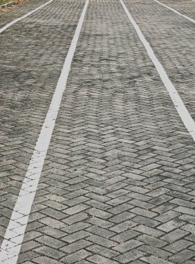 Klasik ringa kemiği iç içe geçmiş bir desen içeren gri tuğlalı kaldırımın temiz ve tepeden tırnağa bir perspektifi. Çerçevenin içinde dikey olarak uzanan iki düz, paralel beyaz çizgi yön ve ölçek hissi yaratıyor..