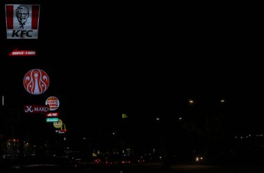 Parlak logolar ve karanlık gökyüzüne karşı perakende satış işaretleri. Sahne, araba ışıkları yolda görünür ve uluslararası ve yerel markaların bir karışımı ile kentsel ticari yaşamı yakalıyor..