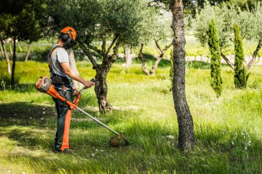 Koruyucu giysiler içinde genç bir adam güneşli bahar gününde bahçede çim biçme makinesiyle yeşil çim biçiyor. Yan görünüm.