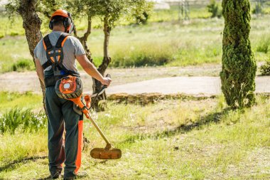 Arka planda, açık hava bahçesinde çimleri budarken kask takan ve koruyucu giysiler giyen bir adam görülüyor..