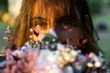 Gün batımında bulanık bir gelin buketinden kameraya dikkatlice bakan genç bir kadının yakın çekimi..