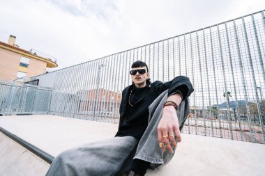 Kişilik kavramları, gurur, Z jenerasyonu. Basket sahasından metal bir çitle ayrılmış kaykay rampasında oturan ve bulutlu bir günde kameraya bakan beyaz olmayan bir yetişkinin alçak açılı portresi..