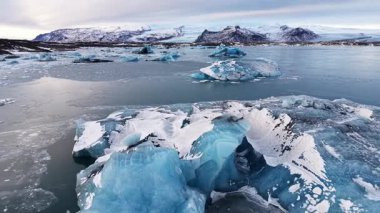 Hava aracı İzlanda 'nın buzul gölünde sürüklenen buzdağlarını geri çekerek ufuktaki karla kaplı dağları, sakin buzul suyunu ve yumuşak gündoğumu yansımalarını gözler önüne seriyor.
