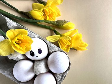 Karton yumurta tepsisinde boyalı gözleri olan, pastel arka planda nergislerle süslenmiş, yazı için yeri olan sevimli bir yumurta. Yüksek kalite fotoğraf. Bahar Kompozisyonu.