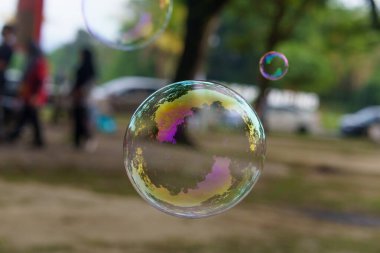 Canlı, yanardöner sabun baloncuklarının yakın plan görüntüsü renkleri yansıtıyor, açık havada insanların ve ağaçların bulanık arka planlarıyla havada süzülüyorlar..