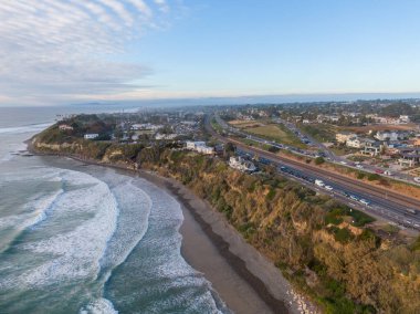 Kaliforniya 'daki Encinitas kıyılarının manzaralı manzarası. Pasifik Okyanusu dalgaları sarp kayalıkların dibindeki kumlu bir plaja yuvarlanıyor. Bu kumsalda yoğun bir sahil yolu ve sakin bir sabah gökyüzü altındaki yerleşim yerlerini destekliyor..