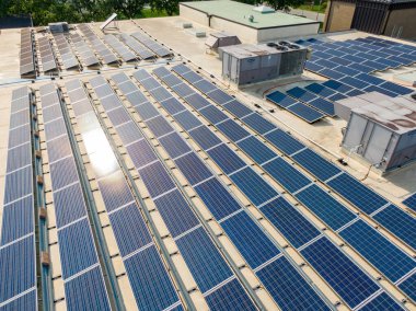 Düz bir ticari çatıdaki devasa bir fotovoltaik güneş dizisinin detaylı hava görüntüsü. Mavi paneller, büyük endüstriyel havalandırma üniteleri ve egzoz deliklerini çevreleyen ve sürdürülebilir inşaat işlemleri için sabah güneşini yakalayan sıralar halinde sıkıca paketlenmiştir..