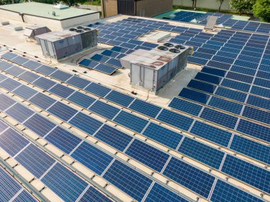 Düz bir ticari çatıdaki devasa bir fotovoltaik güneş dizisinin detaylı hava görüntüsü. Mavi paneller, büyük endüstriyel havalandırma üniteleri ve egzoz deliklerini çevreleyen ve sürdürülebilir inşaat işlemleri için sabah güneşini yakalayan sıralar halinde sıkıca paketlenmiştir..