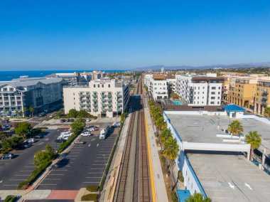Yüksek bir hava manzarası Oceanside şehir merkezindeki tren yolu boyunca kuzeye bakıyor. Sahnede SpringHill Suites Marriott, çatısı havuzlu modern apartman kompleksleri ve berrak mavi gökyüzü altında geçiş platformları yer alıyor..