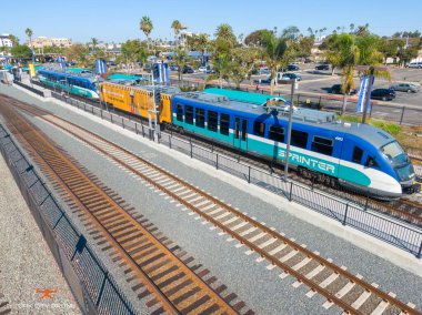 An aerial view of the Oceanside Transit Center featuring a blue and teal SPRINTER hybrid rail train at the platform. Parallel tracks, palm trees, and a parking lot define this Southern California transportation hub under a clear sky.