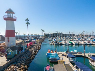Pasifik Okyanusu 'na doğru dar bir liman kanalından bakan bir hava manzarası. Oceanside Liman Köyü 'nün ikonik kırmızı ve beyaz çizgili deniz feneri solda, rıhtım yolunun, hediyelik eşya dükkanlarının ve renkli kanoların yanında duruyor..