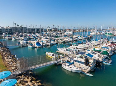 Oceanside Limanı 'nın sakin mavi sularına yanaşmış yelkenli ve motorlu yatları gösteren bir hava manzarası var. Sahnede, arka planda görünen palmiyeli kıyılar ve kıyı tepeleri ile berrak bir gökyüzü altında yoğun direkler yer alıyor..