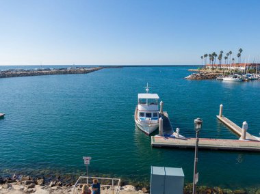 Açık bir günde Oceanside Limanı 'nın ana kanalına bakan bir hava manzarası. Ön planda bir kürekçi kameraya el sallarken, birkaç tekne ve bir yelkenli de kenli yatlar ve palmiye ağaçları arasındaki mavi sularda geziniyor..