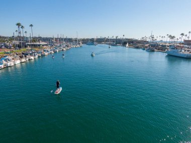 Oceanside Limanı 'nın ana kanalına bakan bir hava manzarası. Ön planda bir kürekçi, kenetlenmiş yelkenliler ve hareket halindeki birkaç küçük motorlu botla çevrili derin mavi sularda gezinirken kameraya el sallıyor..
