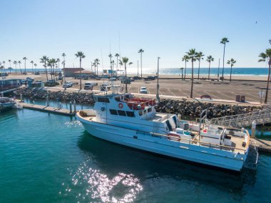 Hava görüntüsü, Oceanside Limanı 'nın parlak turkuaz suyuna kenetlenmiş büyük beyaz bir spor gemisine odaklanıyor. Sahnede kaya kaplı bir iskele, palmiye ağaçları ve Pasifik Okyanusu ufkuna bakan güneşli bir otopark yer alıyor..