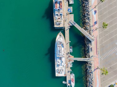 Yüksek açılı bir hava manzarası Oceanside Limanı 'ndaki beton iskeleye kenetlenmiş iki büyük beyaz gemiye bakıyor. Parlak turkuaz su gri park yeriyle ve palmiye ağaçlarıyla kaplı bir kayayla çelişiyor..