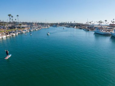Oceanside Limanı 'nın aşağısına bakan bir hava manzarası. Birkaç motorlu tekne derin mavi sularda yüzerken, bir kürekçi soldan dalgalar savuruyor. Yelkenliler her iki taraftaki rıhtımlarda açık ve parlak bir gökyüzünün altında..