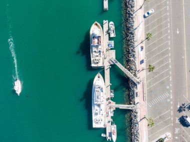 Yukarıdan aşağıya bakan bir görüntü, Oceanside Limanı 'ndaki bir rıhtıma yanaşmış iki büyük beyaz tekne yakalıyor. Küçük bir motorlu tekne canlı turkuaz sularda dolaşır, palmiye ağaçlarıyla dolu bir otoparkın yanında beyaz bir iz bırakır..
