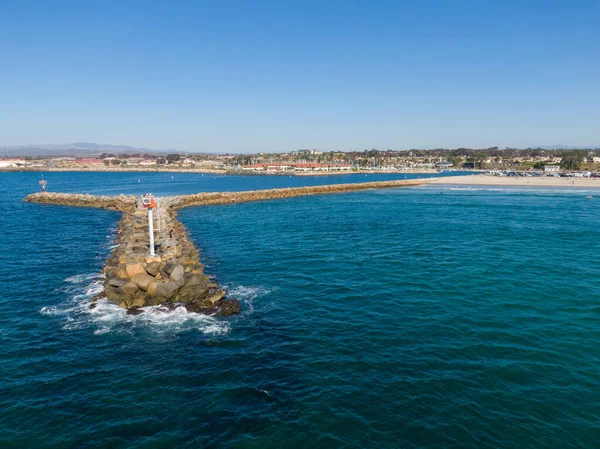 Bir hava manzarası, derin mavi Pasifik Okyanusu 'na uzanan uzun bir kaya iskelesini vurgular. Dalgakıranın sonunda beyaz bir navigasyon ışığı duruyor. Okyanus kıyısının girişini ve açık bir gökyüzünün altındaki komşu kumsalı koruyor..