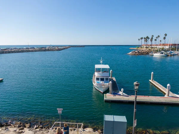 Açık bir günde Oceanside Limanı 'nın ana kanalına bakan bir hava manzarası. Ön planda bir kürekçi kameraya el sallarken, birkaç tekne ve bir yelkenli de kenli yatlar ve palmiye ağaçları arasındaki mavi sularda geziniyor..