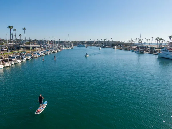 Oceanside Limanı 'nın ana kanalına bakan bir hava manzarası. Ön planda bir kürekçi, kenetlenmiş yelkenliler ve hareket halindeki birkaç küçük motorlu botla çevrili derin mavi sularda gezinirken kameraya el sallıyor..