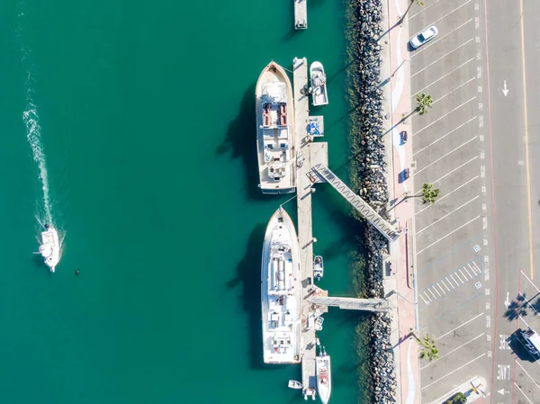 Yukarıdan aşağıya bakan bir görüntü, Oceanside Limanı 'ndaki bir rıhtıma yanaşmış iki büyük beyaz tekne yakalıyor. Küçük bir motorlu tekne canlı turkuaz sularda dolaşır, palmiye ağaçlarıyla dolu bir otoparkın yanında beyaz bir iz bırakır..