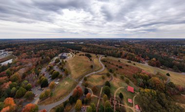 This expansive drone photograph captures the breathtaking autumn landscape of Wake Forest, North Carolina. The dense forest canopy is transformed with brilliant orange and yellow fall foliage surrounding Joyner Park. Perfect for travel and tourism.