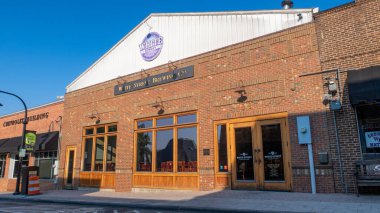 Wake Forest, NC 'deki White Street Brewing Co. binasının sokak seviyesinde çekilmiş bir resmi. Tarihi tuğla mimarisi, geniş ahşap çerçeveli pencereler, beyaz bir çatı çizgisi ve bira fabrikasının imzalı dairesel logosunu berrak bir gökyüzünün altında barındırır..