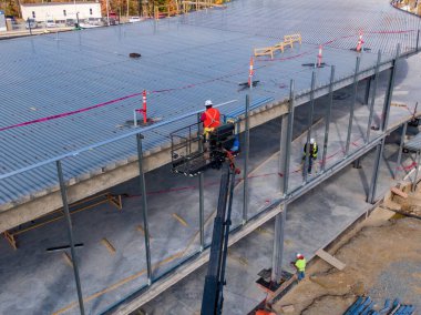 Bir insansız hava aracı modern ticari yapıya geniş çaplı metal çatı kuran işçileri yakalıyor. Sahne endüstriyel mimari, inşaat güvenlik ekipmanları ve büyük bir kalkınma projesinin hassas mühendisliğini vurguluyor.