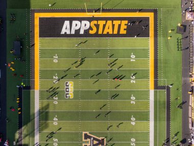 Dik bir İHA atışı Kidd Brewer Stadyumu 'ndaki FieldTurf' a bakıyor. Görüntü, futbolcuların canlı siyah ve altın çim üzerinde konumlanarak, Boone 'daki levhalar ve 