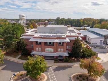 Kuzey Carolina 'daki kırmızı tuğlalı Apex Town Hall' un havadan görünüşü, beyaz kenarlı modern mimari içeriyor. Çekimde arka plandaki beyaz su kulesi, çevre yeşillik ve parlak bir gökyüzünün altında asfalt bir cadde yer alıyor..