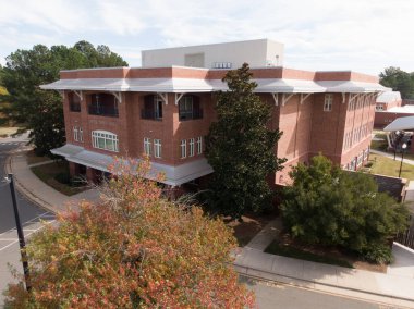 Kuzey Carolina 'daki kırmızı tuğlalı Apex Town Hall' un havadan görünüşü, beyaz kenarlı modern mimari içeriyor. Çekimde arka plandaki beyaz su kulesi, çevre yeşillik ve parlak bir gökyüzünün altında asfalt bir cadde yer alıyor..