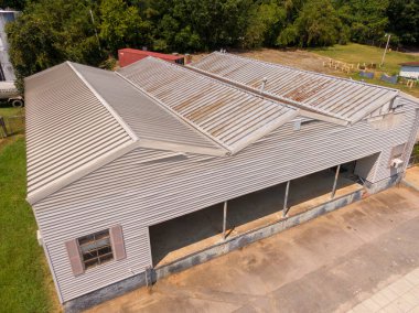 Yüksek açılı hava görüntüsü, endüstriyel bir binadaki yıpranmış metal çatı sistemini gösteriyor. Paneller, bej kaplama ve yoğun yeşil ağaç çizgisi ile sınırlandırılmış çeşitli çıkıntılı bölgelerde yüzey paslanması ve oksitlenme sergiliyor..