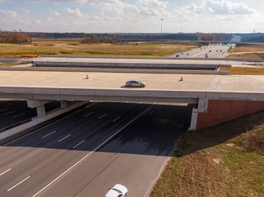 Bu hava görüntüsü, Kuzey Carolina, Raleigh yakınlarındaki 40 numaralı otoban üzerinden geçen gümüş rengi bir arabayı gösteriyor. Sahne modern beton altyapıyı, otoyol inşaat konilerini ve sonbahar alanlarını yakalıyor..