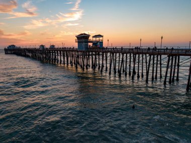 Düşük açılı bir insansız hava aracı Pasifik Okyanusu 'na uzanan Oceanside İskelesi' nin ahşap kazıklarını yakalıyor. Batan güneş, suyun yüzeyinde altın bir parıltı yaratırken insanların siluetleri, yumuşak bir akşam gökyüzünün altında yapı boyunca yürür..