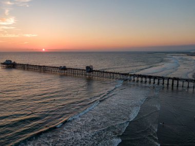 Pasifik Okyanusu 'nun üzerinde güneş batarken tarihi Okyanus Kıyısı' nın geniş bir hava manzarası. Altın güneş ufukta asılı, dalgaların arasında parıldayan bir ışık izi bırakıyor ve kumlu sahilde az sayıda insan yürüyor..