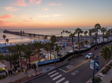 Oceanside, California 'nın yükseltilmiş manzarası, günbatımında Pasifik Okyanusu' na uzanan tarihi iskeleyi gösteriyor. Plaj bölgesinde renkli çadırlar ve palmiye ağaçları hizalanırken, bir araba canlı bir akşam gökyüzünün altında kıyı yolu boyunca ilerliyor..