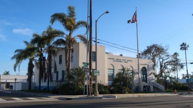 Oceanside, California 'daki Birleşik Devletler Postanesinde açık mavi gökyüzü altında klasik Art Deco mimarisi sergileniyor. Amerikan ve savaş esirleri palmiye ağaçlarının yanında dalgalanırken, bu güneşli Güney Kaliforniya sahnesini sokak tabelaları ve bir posta kutusu tamamlıyor..