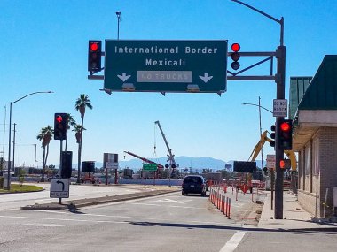 Büyük yeşil bir otoyol tabelası trafiği Mexicali 'deki Uluslararası Sınır' a yönlendiriyor. Sahne trafik ışıkları, palmiye ağaçları ve inşaat vinçleriyle dolu yoğun bir kavşağı gösteriyor..