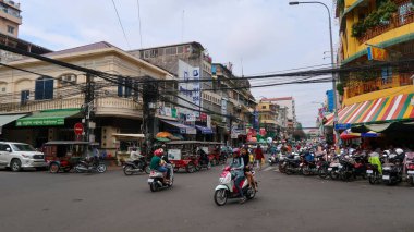 Phnom Penh 'de hareketli bir sokak köşesinde kaotik bir elektrik hattı ağı bulunuyor. Motosikletçiler ve tuk-tuklar kavşakta dolaşırken, çizgili güneşlikleri ve park etmiş scooterları olan sarı bir bina kentin canlı kentsel yaşamını ön plana çıkarıyor..