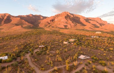 Altın Kanyon 'daki Batıl inanç Dağları' nın havadan görünüşü, AZ, altın saat. Sıcak ışık, saguaro kaktüsü ve kırsal evlerle dolu bir çöl arazisinin üzerindeki engebeli tepeleri vurur. Yumuşak, parlak günbatımı gökyüzünün altında manzaralı bir Sonoran Çöl manzarası.