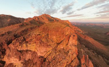 Altın Kanyon, AZ 'deki güneşten sırılsıklam olmuş Batıl inanç Dağları' nın insansız hava aracı görüntüsü. Altın saat ışığı engebeli volkanik kaya yüzeylerinde ve çöl sırtlarında parlar. Uçsuz bucaksız Sonoran Çölü 'nün üzerindeki devasa uçurum kulesi yumuşak, boyalı günbatımı gökyüzünün altında..
