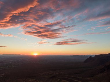 Arizona çölünün üzerinde nefes kesen gün batımı. Güneş ufkun altına batarken, derin turuncu ve pembe bulutlar gökyüzüne doğru uzanıyor, yayılan vadi ve uzak dağ siluetleri üzerine sıcak bir ışıltı saçıyor. Sakin güneybatı akşamı.