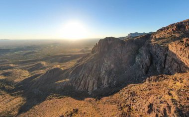 Arizona 'daki Altın Kanyon' daki Batıl inanç Dağları 'nda güneşin batışını gösteren hava aracı görüntüsü. Ufukta parlak güneş ışıkları parlıyor, engebeli çöl uçurumlarını aydınlatıyor ve Sonora 'nın engin manzarasını sıcak, altın saatli bir parıltıyla aydınlatıyor..
