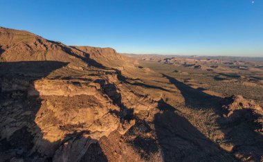 Wide aerial drone view of the Superstition Mountains in Gold Canyon, Arizona, during the late afternoon. Jagged volcanic cliffs cast long, dramatic shadows across the rugged Sonoran Desert floor under a clear, pale blue sky at golden hour.